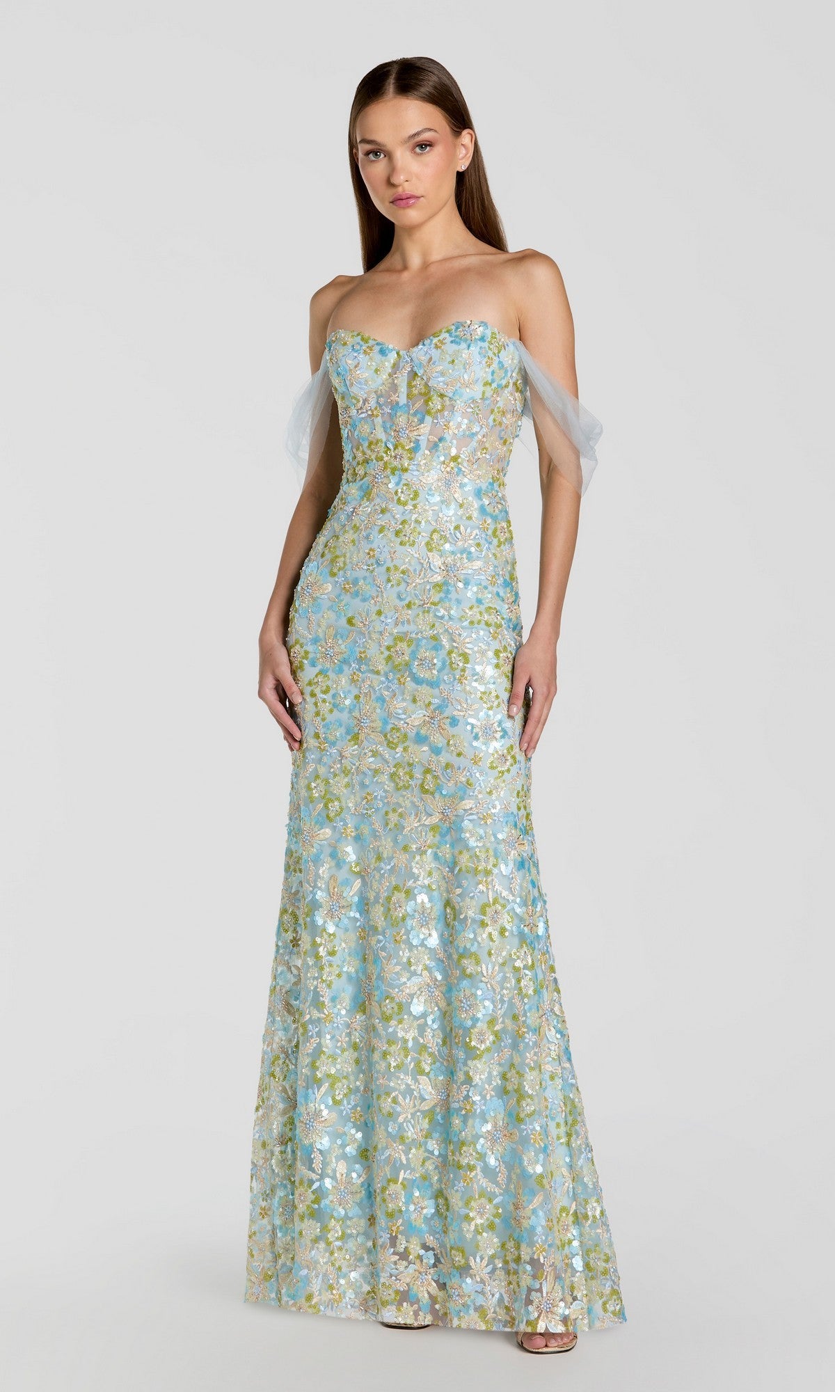 A woman stands against a plain light gray background wearing the Mac Duggal 69045 Long Formal Dress in powder blue, featuring floor-length design, off-the-shoulder tulle straps, and delicate floral embroidery.